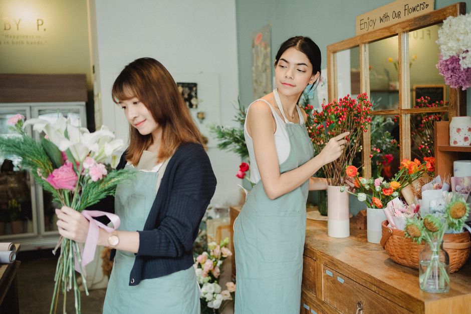 Two women in a flower shop are handling fresh floral arrangements. The woman on the left, with shoulder-length brown hair, holds a bouquet of white lilies, pink carnations, and greenery, wrapped with 