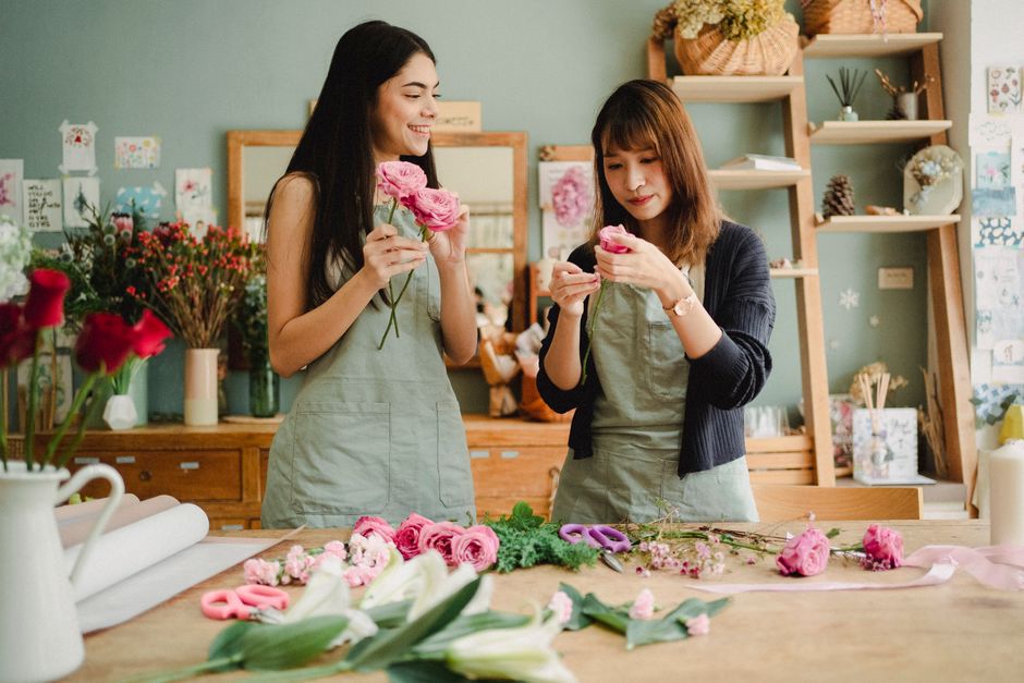 Two women in a floral workshop are holding and examining pink roses, with arrangements of fresh flowers, including lilies and roses, laid out on a wooden table in front of them. One woman has long dar