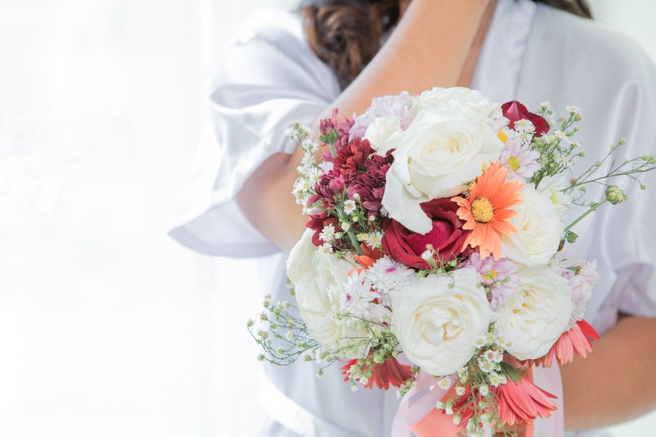 A person holding a floral bouquet arrangement featuring large white roses, red ranunculus, peach daisy-like flowers, and delicate white filler blooms, with green foliage and tiny white accents. The bo