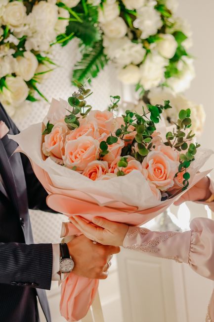 A floral gift featuring a bouquet of soft pink roses with lush, green eucalyptus leaves, wrapped in blush-colored tissue paper and cream-colored wrapping paper, being handed over by a person in cream 