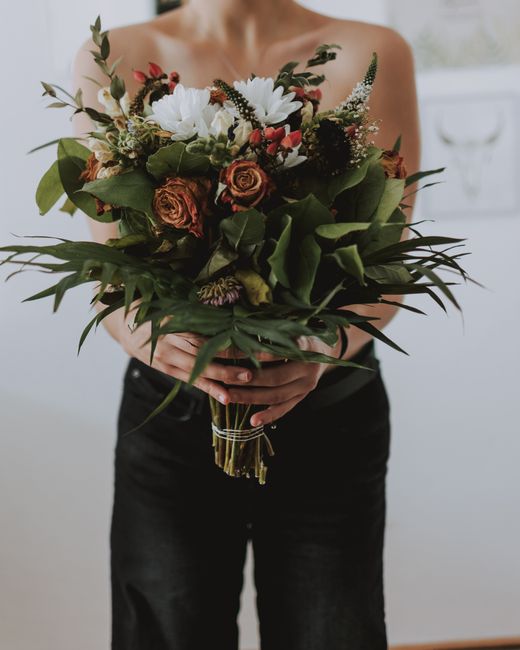 A person holds a large, vibrantly arranged bouquet of fresh flowers, featuring white daisies, small coral roses, sprigs of lavender, and red berries, all surrounded by lush green foliage and dark leaf