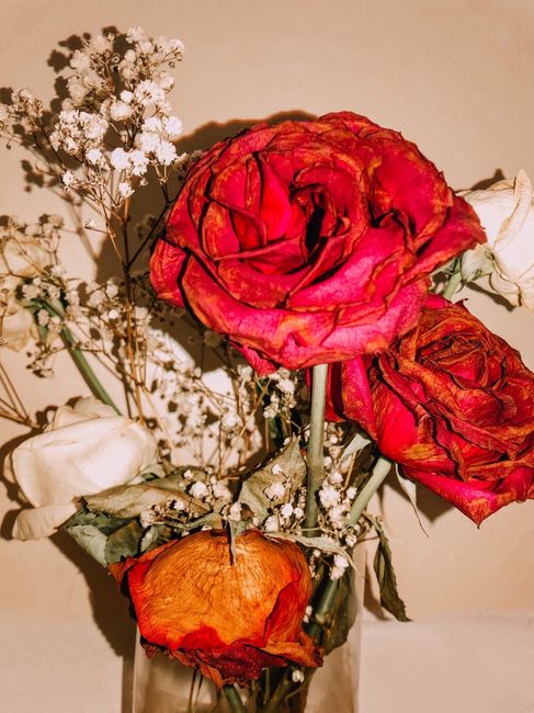 A floral arrangement in a clear glass vase featuring a mix of dried and preserved flowers. The bouquet includes prominent large pink and red roses with a slightly wilted appearance, alongside smaller 