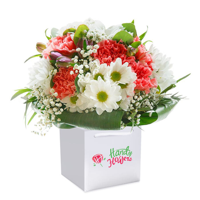 A beautifully designed floral arrangement occupies pride of place inside a clean white gift bag, its charm ready to delight anyone in Rush Green. This lush bouquet combines coral-hued and peach carnations-each petal delicately ruffled-interwoven with clean white daisy-like chrysanthemums, their centers a fresh lime green that catches the eye. Airy sprays of white baby's breath, or gypsophila, fill the spaces between, creating an overall sense of lightness and gentle movement. Here and there, closed buds brushed with hints of purple or mauve, perhaps from alstroemeria, add intriguing depth and promise. Everything is encased in a verdant halo of deep green, glossy leaves, forming a natural collar that anchors the ensemble and echoes the abundant greenery found in local parks like Raphael Park. The pristine white bag, designed for convenient carrying, is fitted with elegant white ribbon handles, a subtle nod to the blend of tradition and modernity found throughout Rush Green. Set against a seamless white background, the bouquet is visually highlighted, celebrating each vivid hue and tender texture with clarity. This composition exudes a sense of ready-to-gift elegance-perfect for celebrations, thoughtful gestures, or commemorating special occasions within the Rush Green community. It embodies the spirit of local flower delivery, offering freshness and heartfelt beauty prepared with expert care by florists who know and cherish the area.